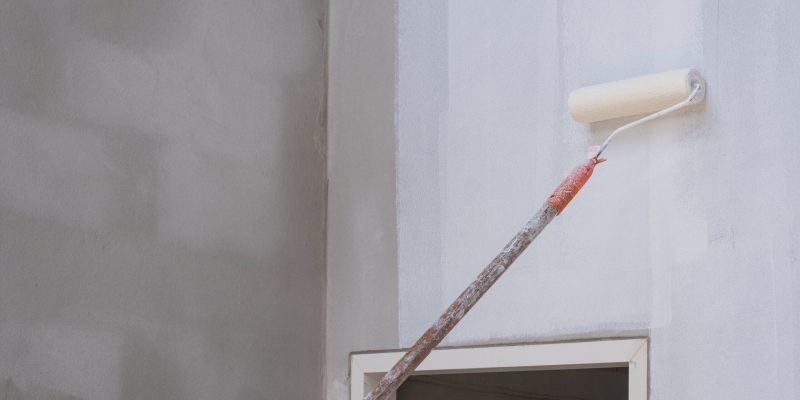 Long handle roller brush applying primer white paint with door frame on cement wall inside of house construction site, building and home renovation concept